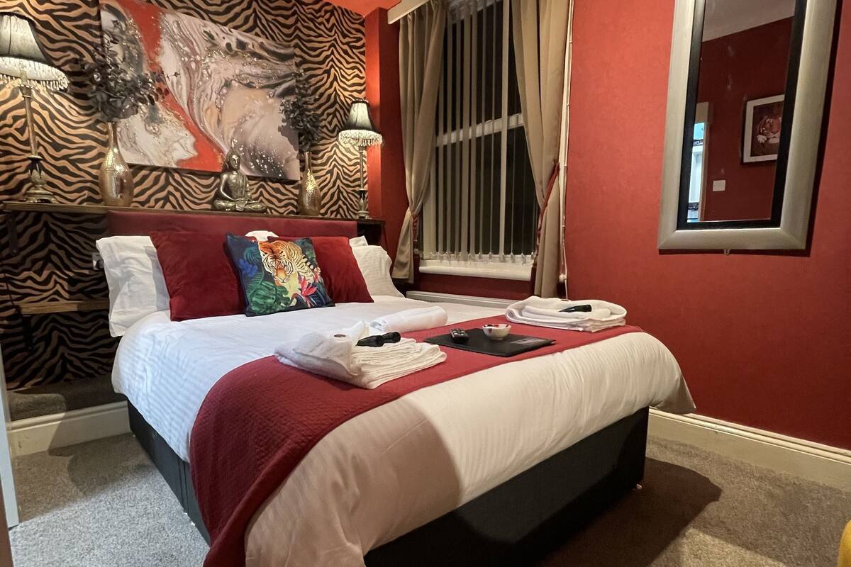 A hotel room with a bed, red walls, a zebra-patterned headboard, a large abstract painting, and a window with curtains.
