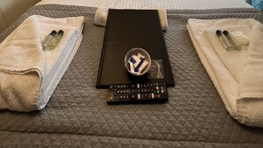 A neatly made bed with a quilted cover, two towels, a remote control, and a small bowl with toiletries. - Delovely Hotel (Blackpool)