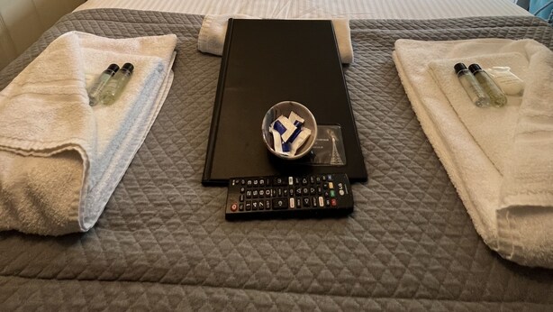 A neatly made bed with a quilted cover, two towels, a remote control, and a small bowl with toiletries. - Delovely Hotel (Blackpool)