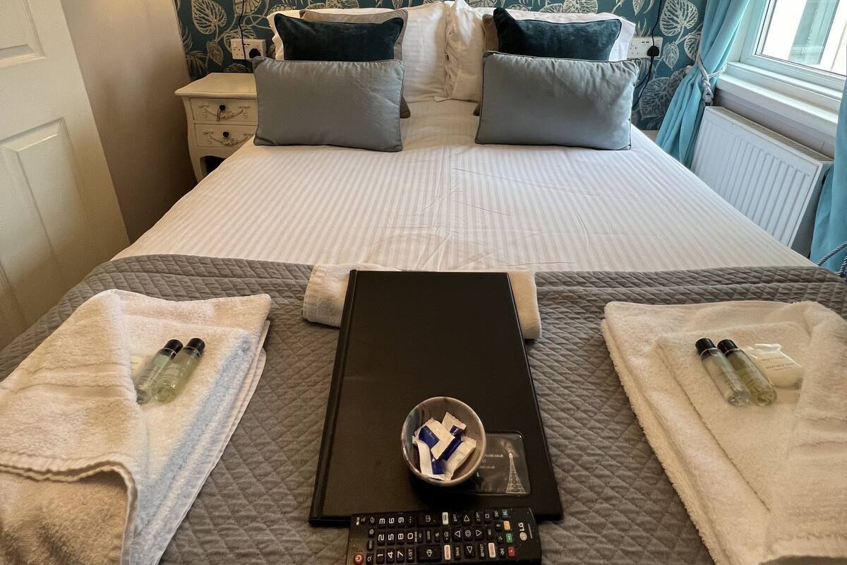 A neatly made bed with a quilted cover, two towels, a remote control, and a small bowl with toiletries.