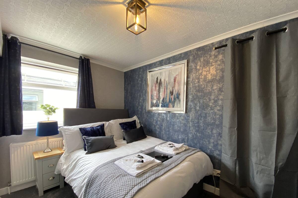 A neatly arranged bedroom with a bed, bedside tables, a radiator, a window with curtains, a painting on the wall, and a ceiling light fixture.