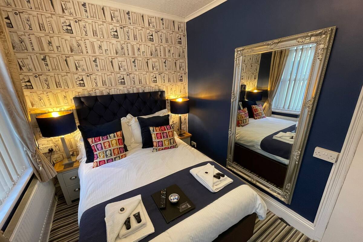 A neatly made bed with a black headboard, white bedding, and colorful pillows. There is a large mirror on the wall and a wall-mounted lamp beside the bed.