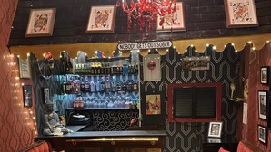 A bar area with a sign reading "Nobody gets out sober", a chandelier, and a bar stocked with various bottles. There are red upholstered seating areas and a striped carpet. - Delovely Hotel (Blackpool)