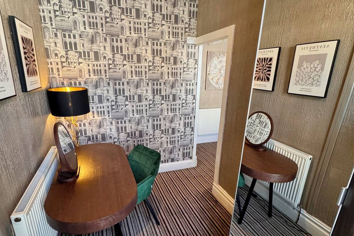 A room with a patterned wallpaper, a round wooden table, a green chair, a small round table, and a mirror on the wall.