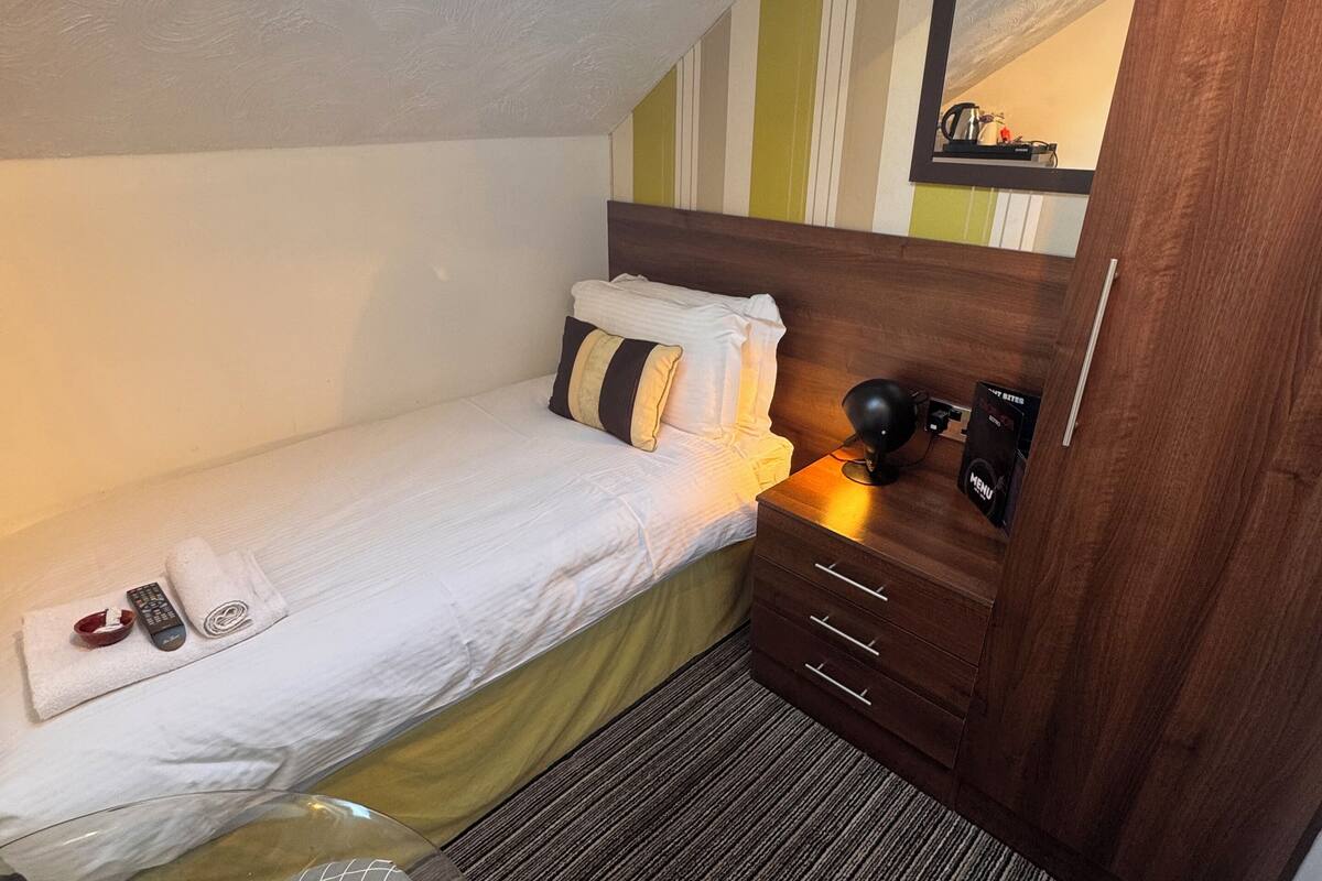 A compact hotel room with a single bed, a bedside table with a lamp, a small round seating area, and a mirror on the wall.