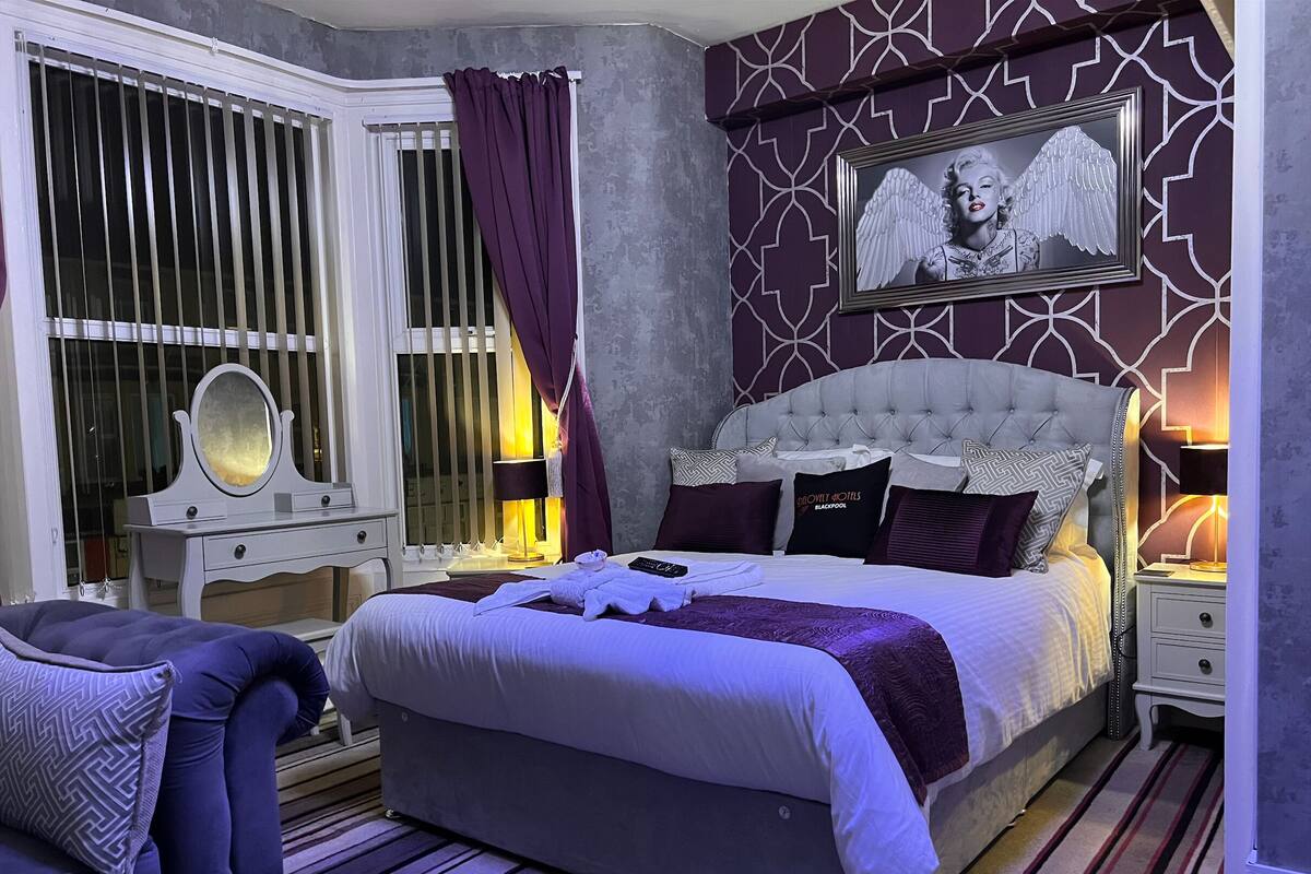 A bedroom with a large bed, a vanity with a mirror, a chair, and a wall with a framed picture.