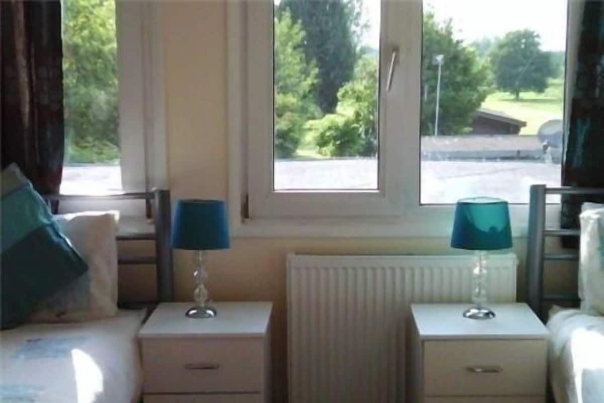A bedroom with a bed, two bedside tables with lamps, a radiator, and a window with a view of greenery outside.