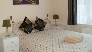 A bedroom with a bed, two nightstands, two lamps, a window with curtains, and a painting on the wall. - London Heathrow Guesthouse (Slough)