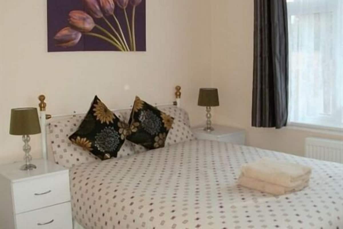 A bedroom with a bed, two nightstands, two lamps, a window with curtains, and a painting on the wall.