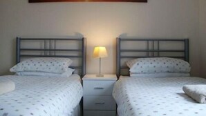 A room with two beds, each with a metal headboard and white bedding with a dotted pattern. There is a nightstand with a lamp between the beds, and a painting of a sunset on the wall above the beds. - London Heathrow Guesthouse (Slough)