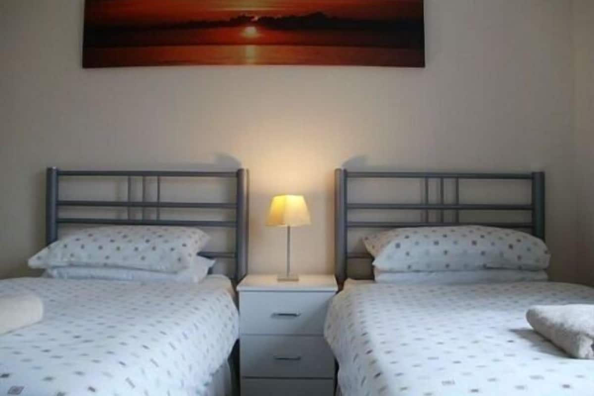 A room with two beds, each with a metal headboard and white bedding with a dotted pattern. There is a nightstand with a lamp between the beds, and a painting of a sunset on the wall above the beds.