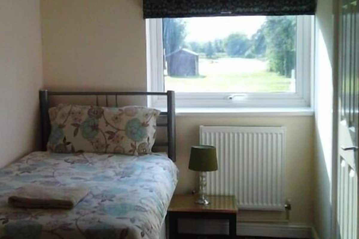 A bedroom with a bed, a nightstand, a lamp, a window with a view of the outdoors, and a radiator.