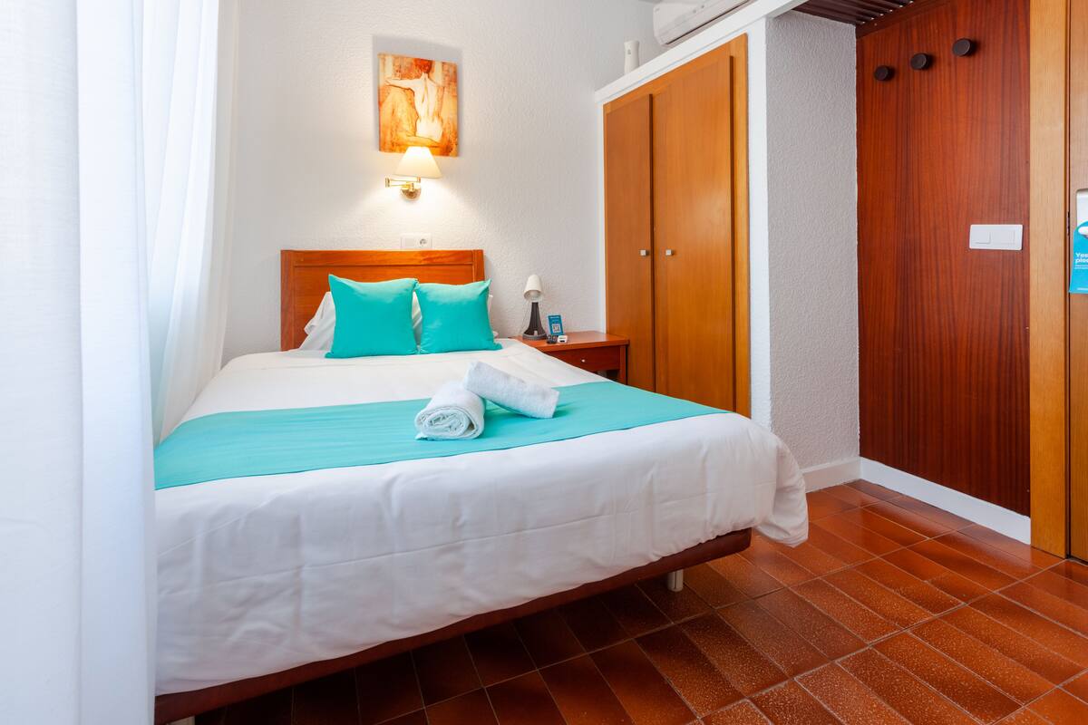 A hotel room with a bed, a nightstand, a wall-mounted lamp, a wooden wardrobe, and a tiled floor.