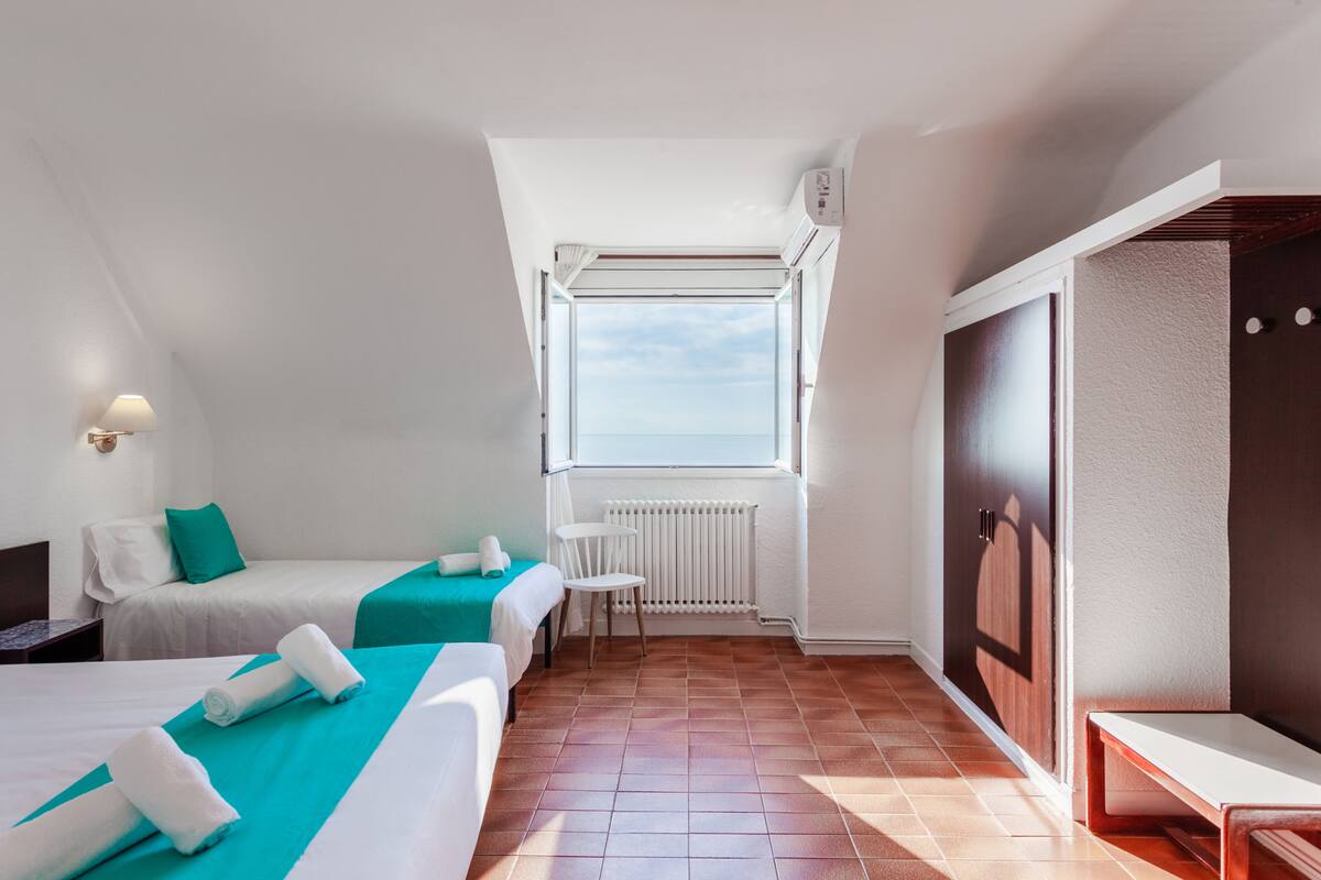 A hotel room with two beds, a small table, a chair, a radiator, and a window with a view.