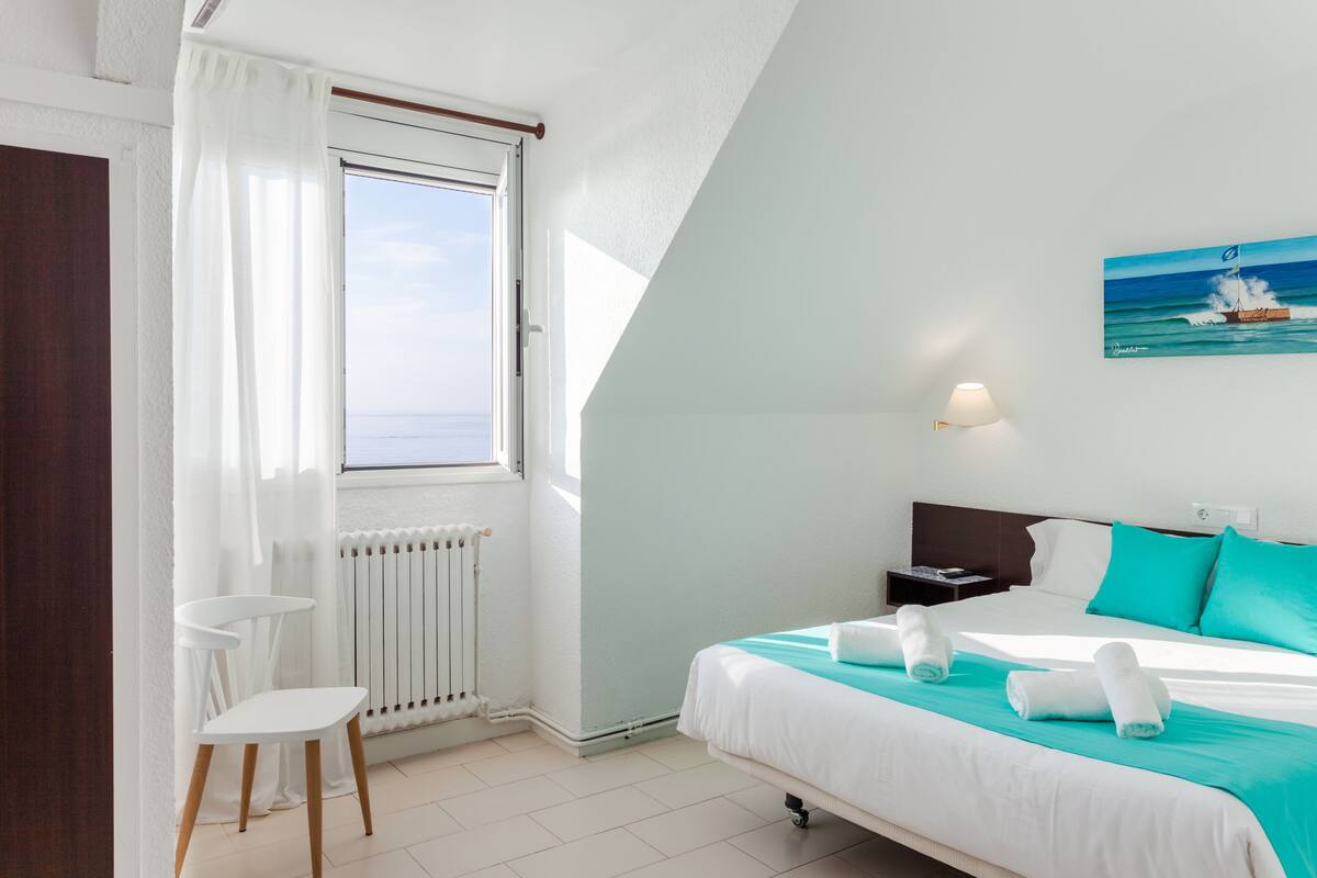 A bedroom with a bed, a window with curtains, a nightstand, and a painting of a boat on the wall.
