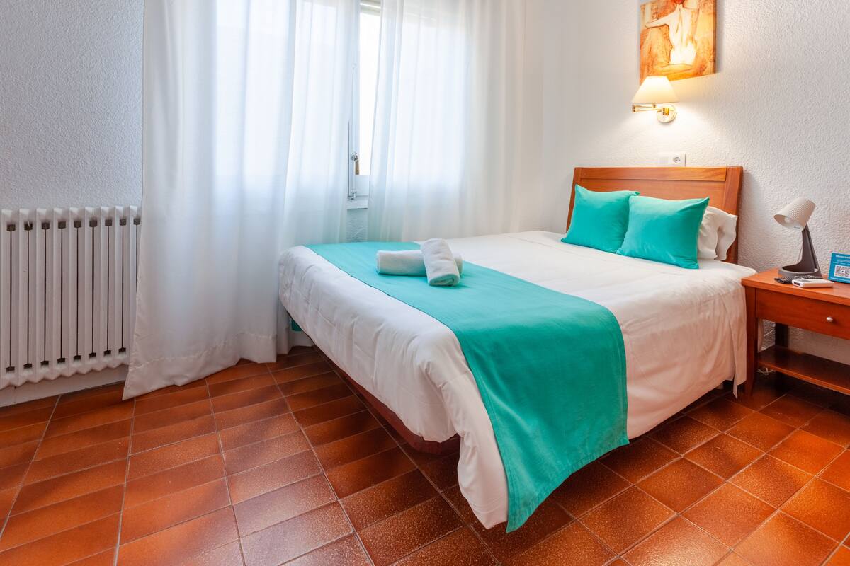A hotel room with a bed, a nightstand, a lamp, a window with curtains, and a radiator.