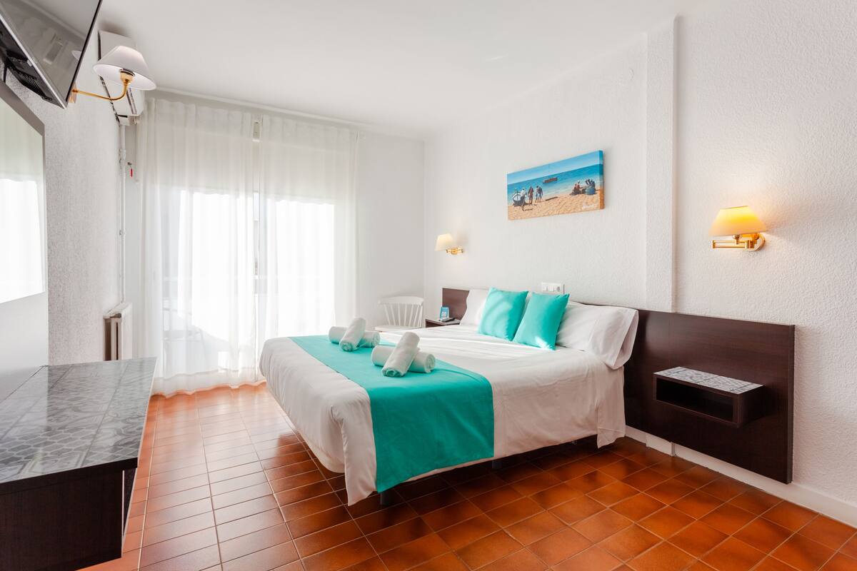 A hotel room with a bed, a desk, a chair, a painting, and a wall-mounted shelf.