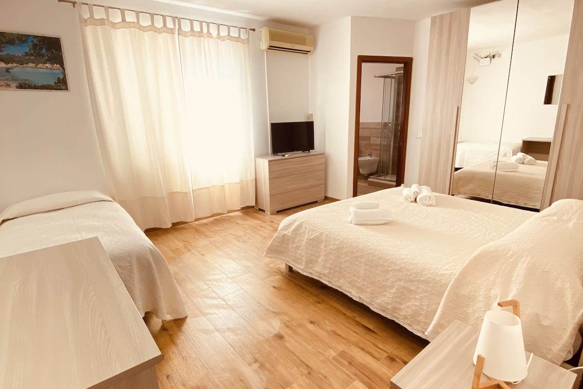 A hotel room with two beds, a wooden floor, a TV, a mirror, and a bathroom visible through an open door.