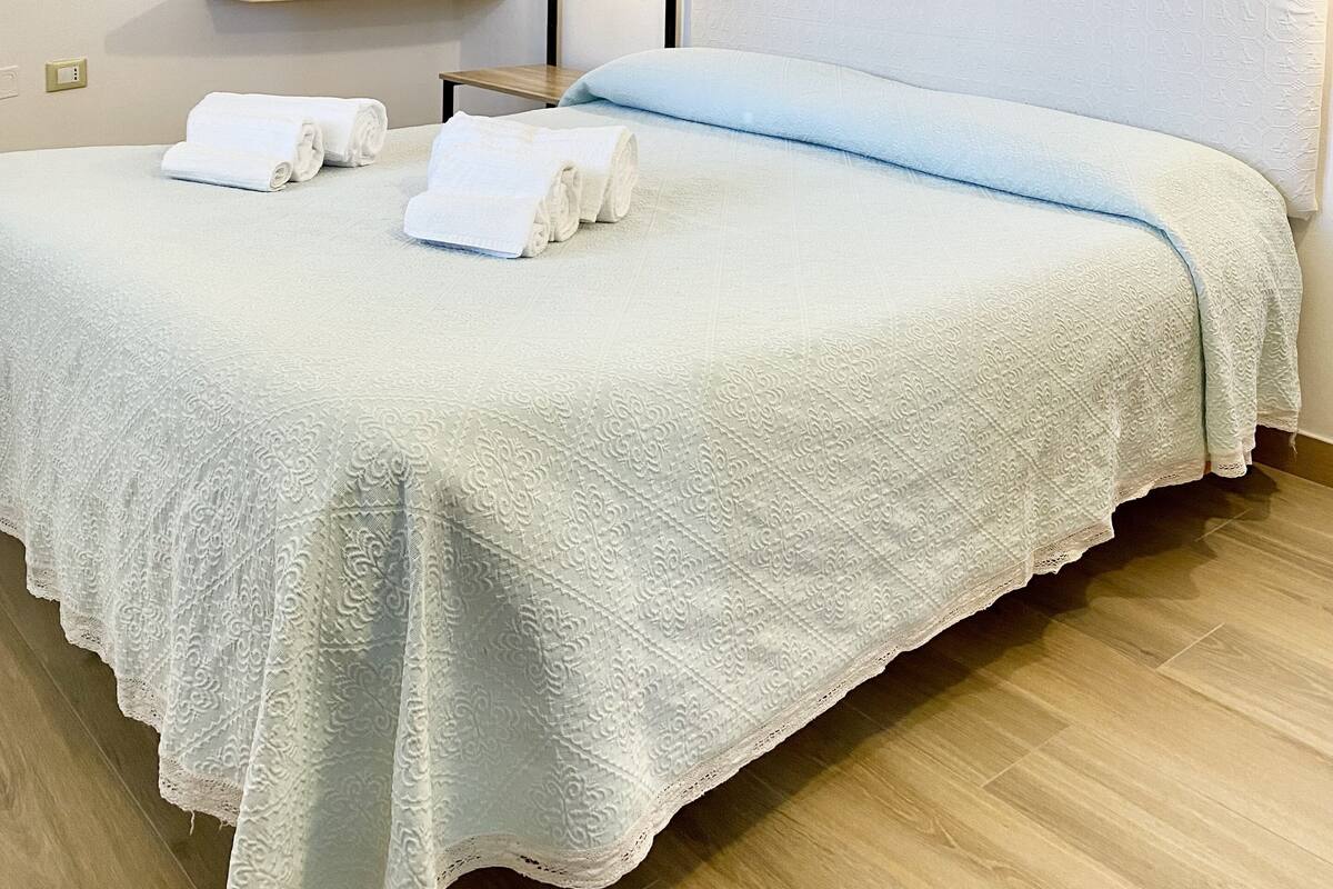 A neatly made bed with white linens and a blue accent, wooden shelves, a mirror, and a wall-mounted light fixture.