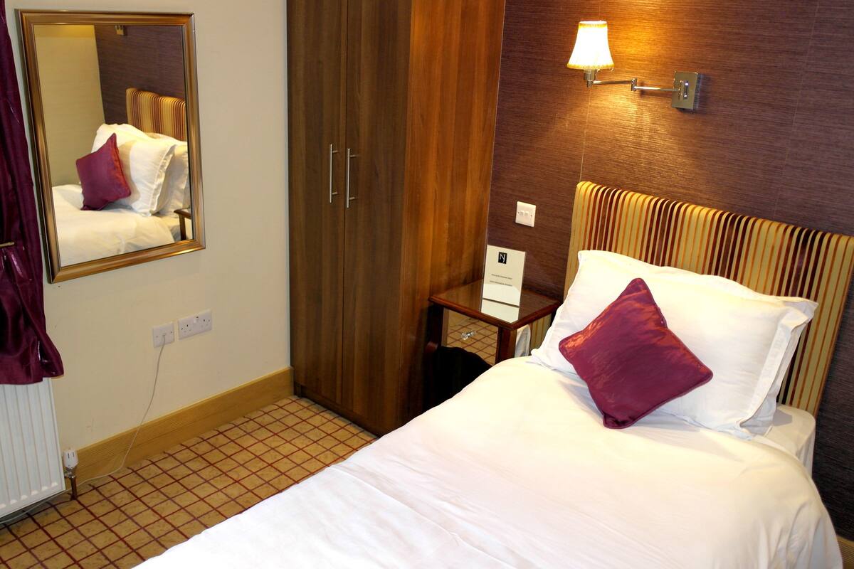 A hotel room with a bed, a mirror, a nightstand, a lamp, and a wardrobe.