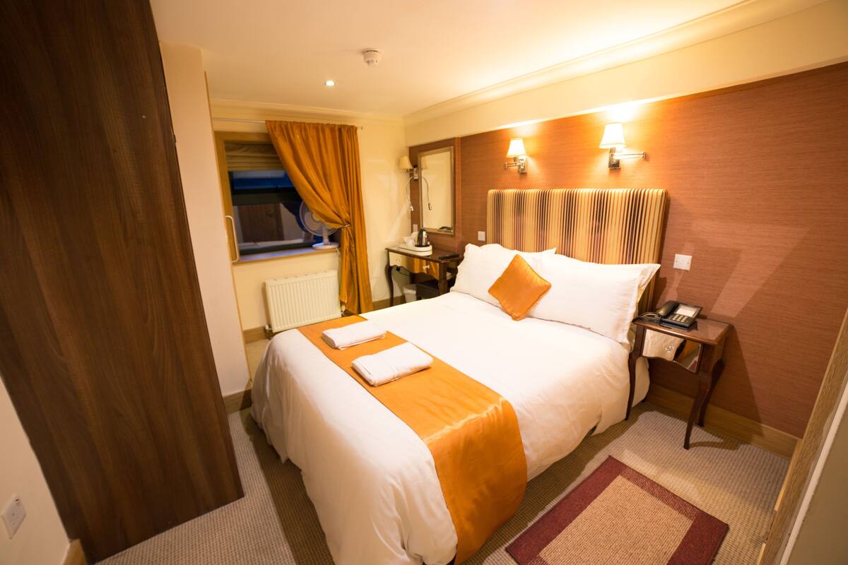 A hotel room with a large bed, a desk with a chair, a nightstand with a lamp, and a window with curtains.