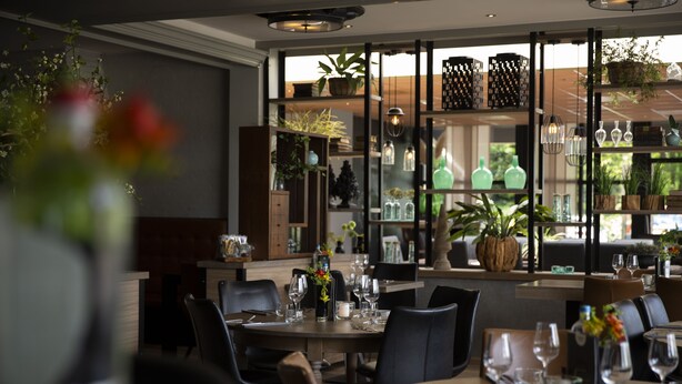 Interior de restaurante con mesas dispuestas para comer, estantes con cristalería y plantas en macetas.