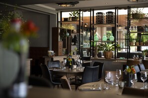 A restaurant interior with tables set for dining, shelves with glassware, and potted plants.