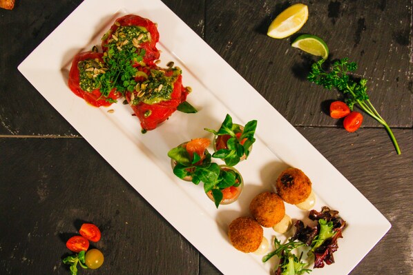 Un plato rectangular blanco con diferentes alimentos, incluyendo tomates rellenos, una ensalada y algunas porciones fritas redondas, sobre una superficie de madera oscura.