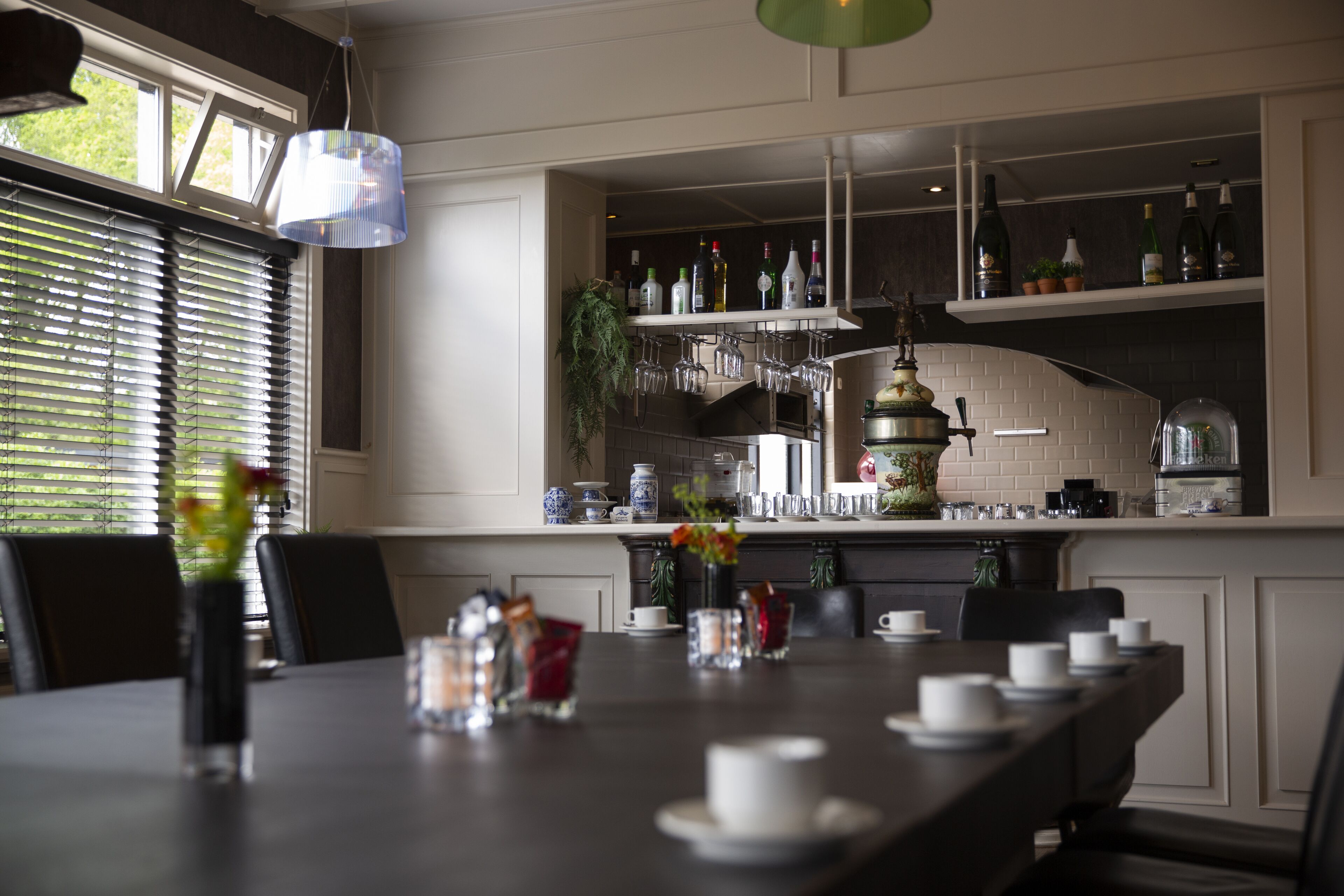 Un comedor con una mesa larga dispuesta para una comida, una zona de bar con estantes llenos de botellas, y vistas al exterior a través de amplias ventanas.