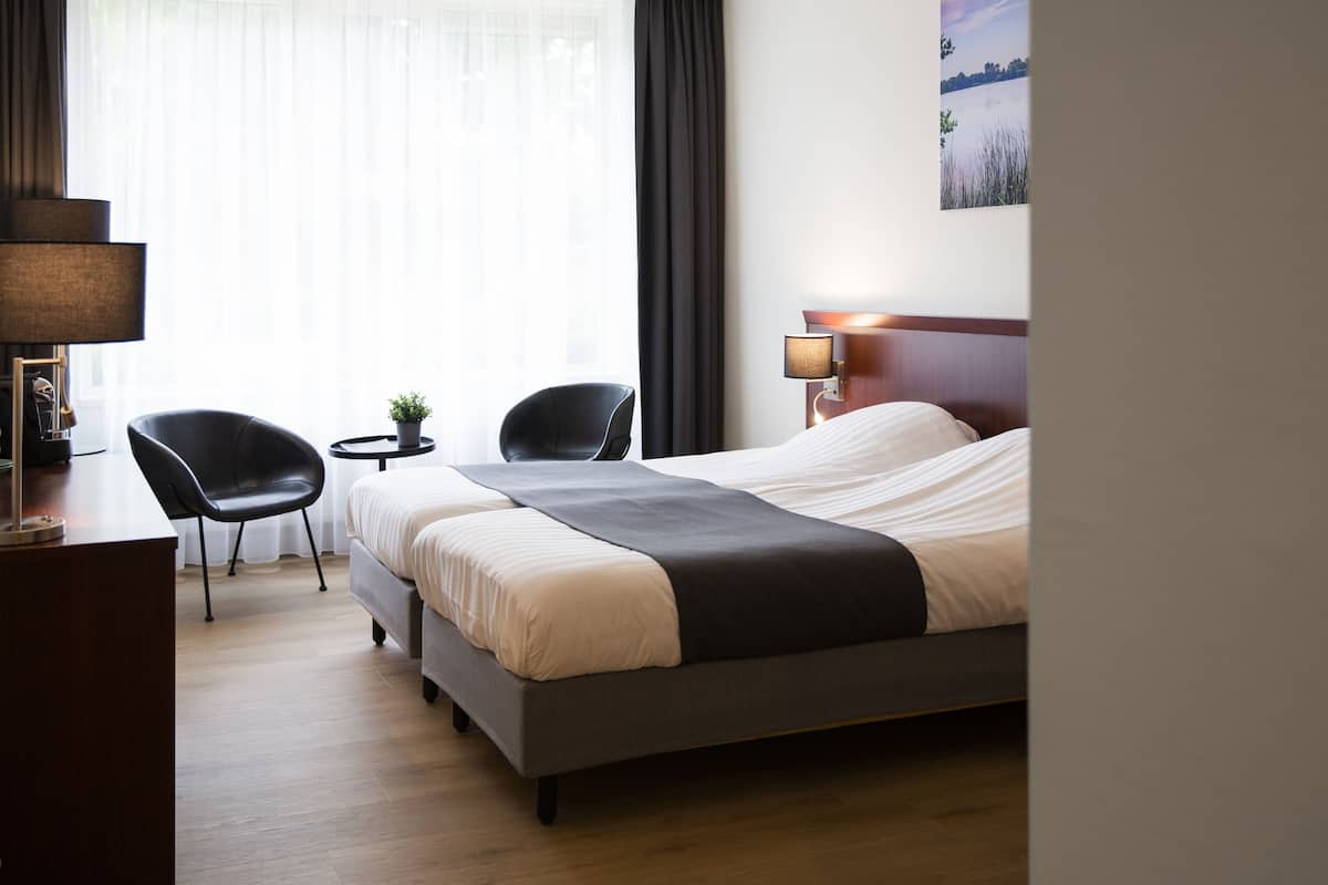 A hotel room with a bed, two chairs, a desk, a lamp, and a painting on the wall.
