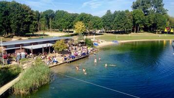 A lakeside swimming area with a dock, people swimming and sunbathing, and a covered seating area.