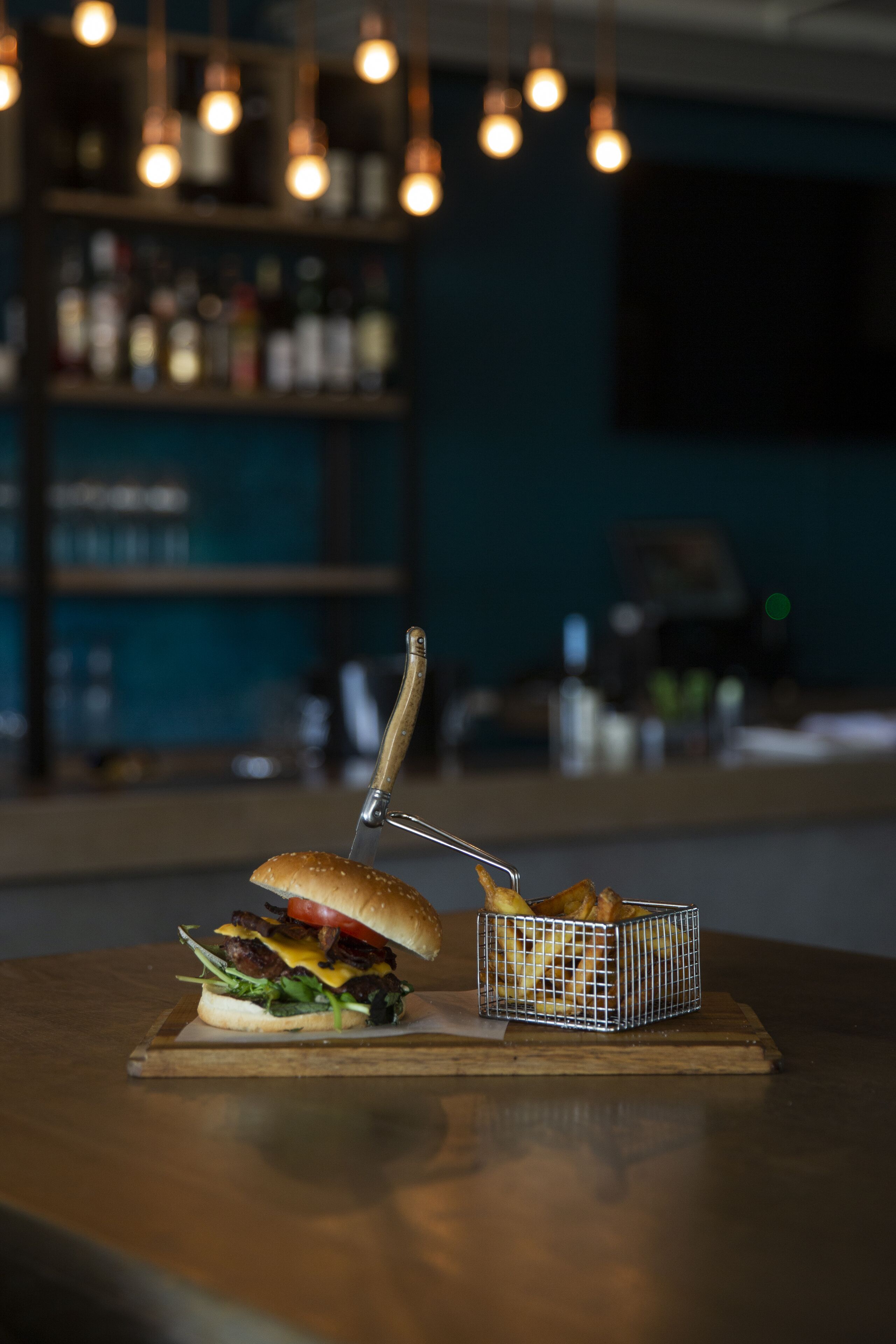A burger on a wooden board with a knife and a basket of fries on a table.