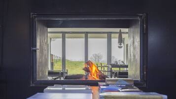 Una habitación con un ventanal grande con vistas a un área verde, una chimenea con leña, y una mesa con libros.