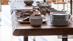 Een eettafel gedekt met borden, kommen en bestek, met uitzicht op bomen en open velden door grote ramen.