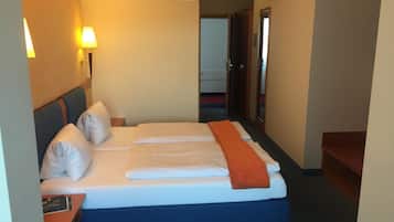 Habitación de hotel con cama, almohadas, mesita de noche, lámpara y puerta que da a otra habitación.