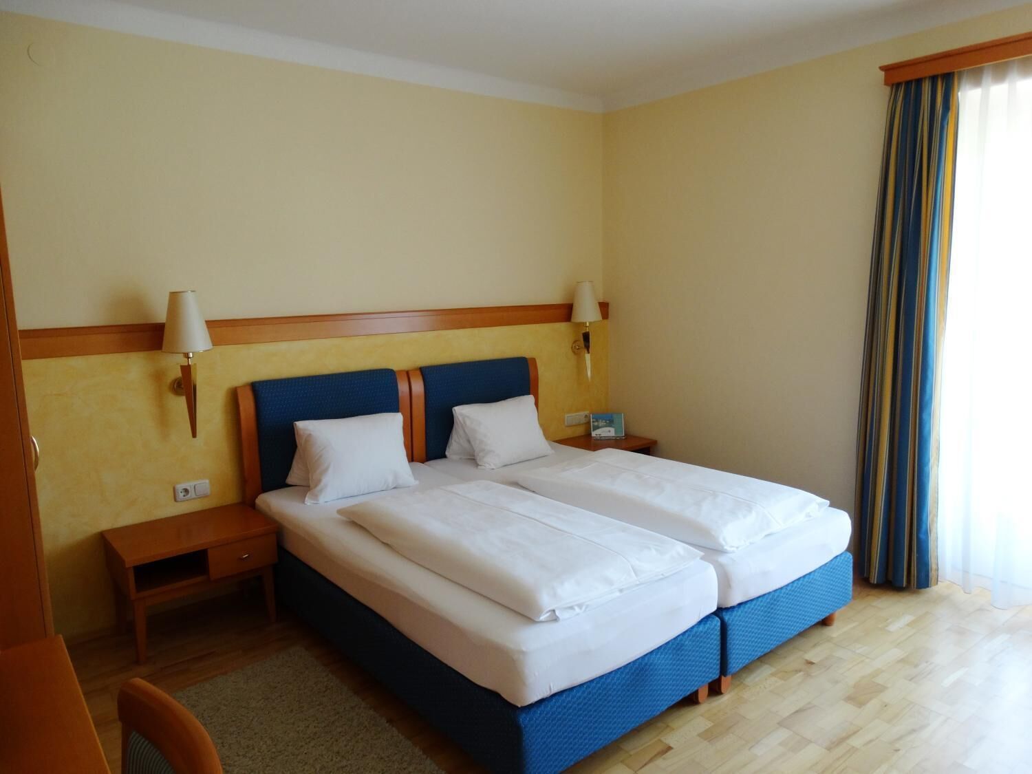 A hotel room with a double bed, two bedside tables, a nightstand, a lamp, and a window with curtains.