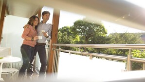 A couple standing on a balcony with a railing, holding cups, and a table with chairs in the background.