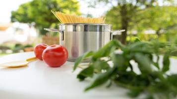 A stainless steel pot with spaghetti on a white surface, accompanied by two tomatoes and a wooden spoon.