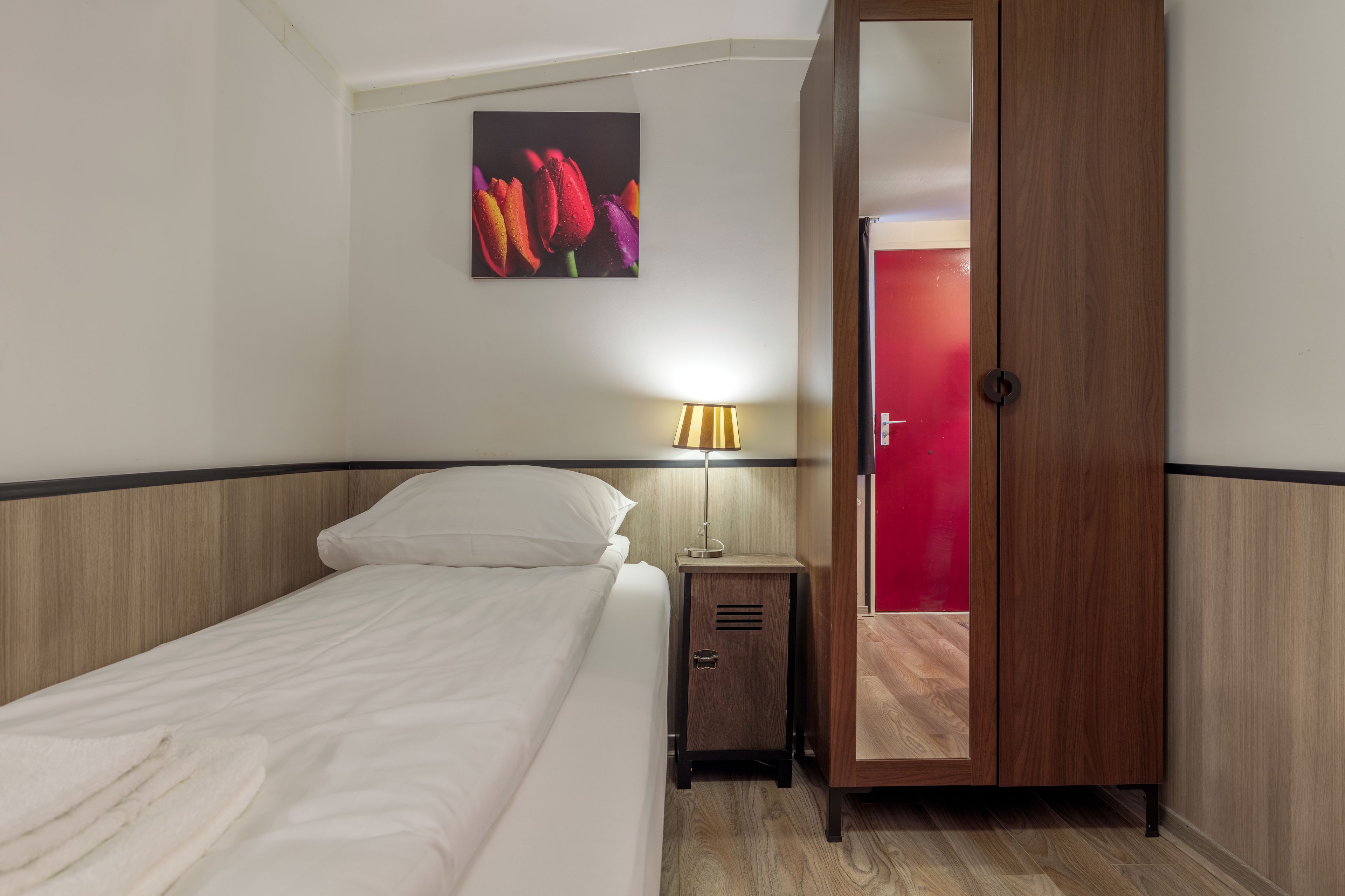 A hotel room with a bed, a nightstand with a lamp, a wardrobe, and a painting of flowers on the wall.