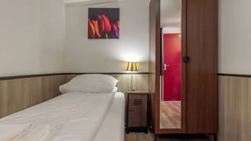 A hotel room with a bed, a nightstand with a lamp, a wardrobe, and a painting of flowers on the wall.