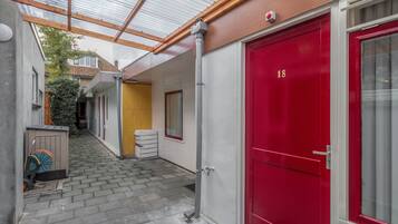En smal korridor med en röd dörr märkt med siffran ’18’, ett överbyggt område med ett tak av metall och ett litet förråd.