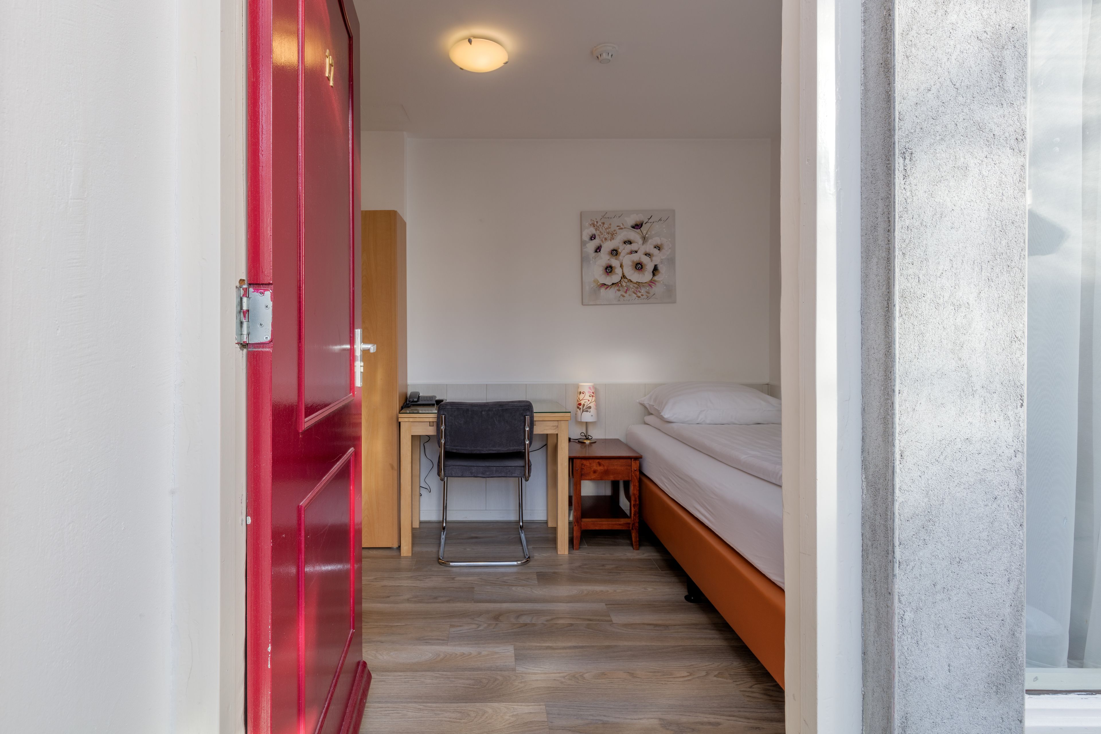 A room with a bed, a desk, a chair, and a red door.