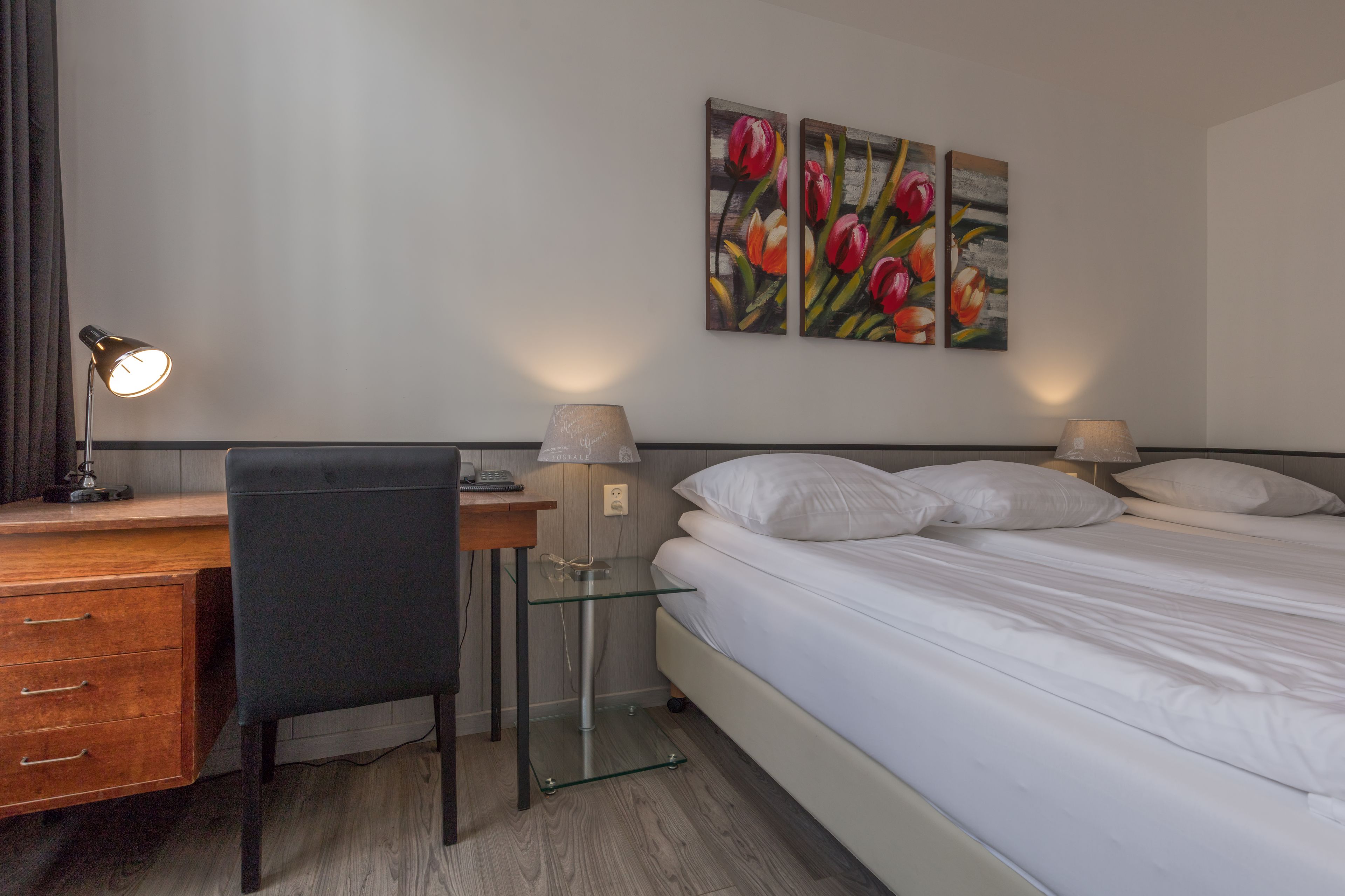 Ein Hotelzimmer mit Bett, Schreibtisch mit Stuhl, einer Lampe und zwei Bildern an der Wand.