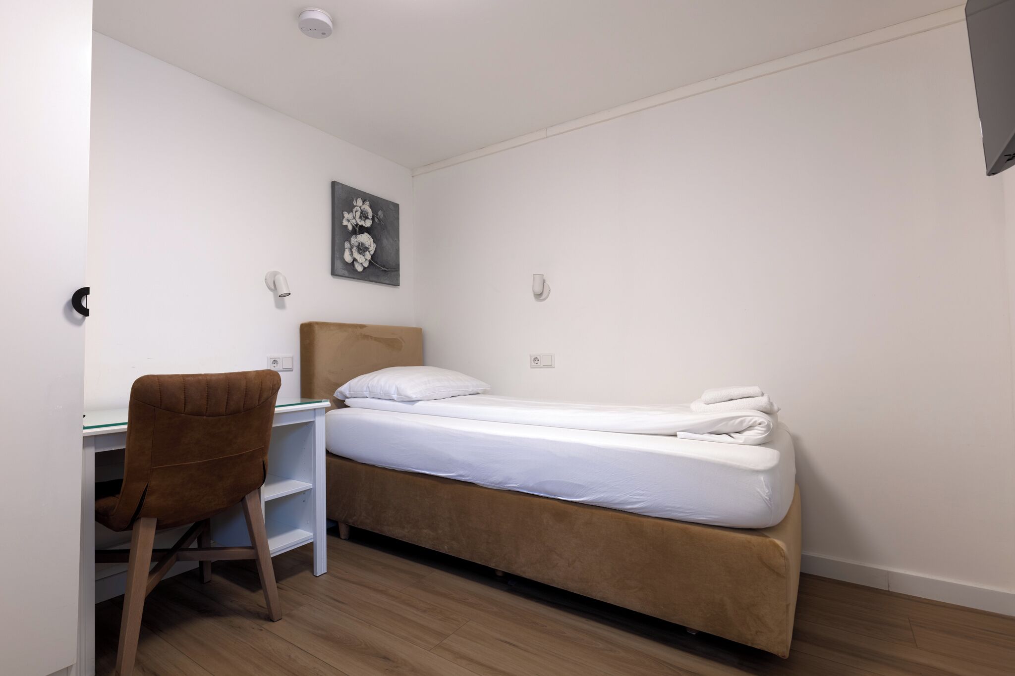 A single bed with a wooden headboard, a small desk with a chair, and a wall-mounted light fixture.
