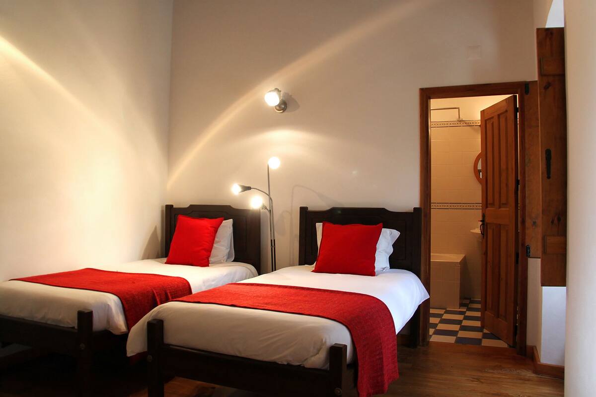 Habitación de hotel con dos camas individuales, cada una con almohada roja y ropa de cama blanca. Se observan lámparas de noche y la puerta del baño.