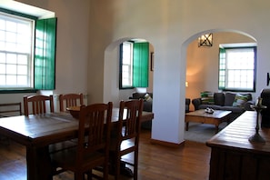 A dining area with wooden tables and chairs, a sofa, and a coffee table. There are windows with green shutters and a lamp on the wall. - Quinta dos Ribeiros (Nisa)