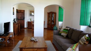 Una sala de estar con una mesa de centro de madera, un sofá con cojines a rayas y un televisor sobre un mueble de madera. Se ven armarios de madera y una puerta verde al fondo.