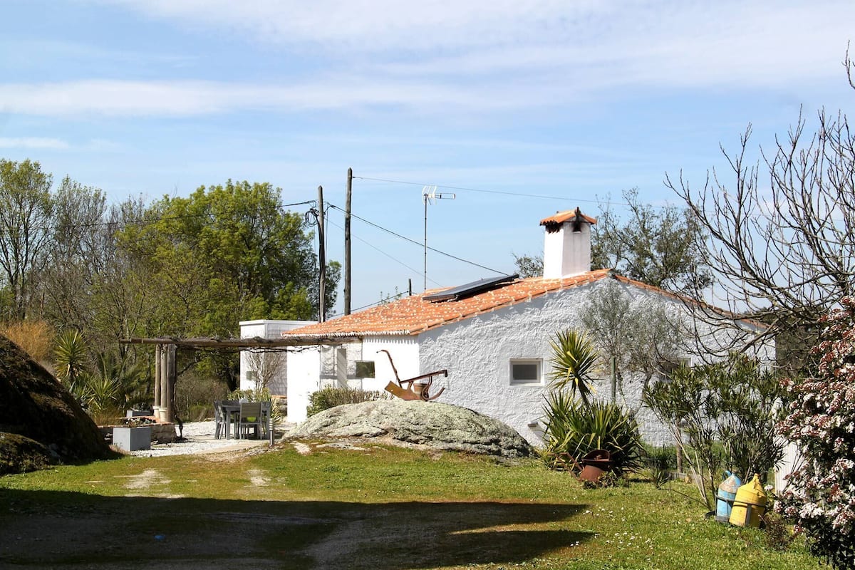 Una casa rústica con techo de tejas, chimenea de piedra y un pequeño jardín con un sendero que lleva a la entrada.