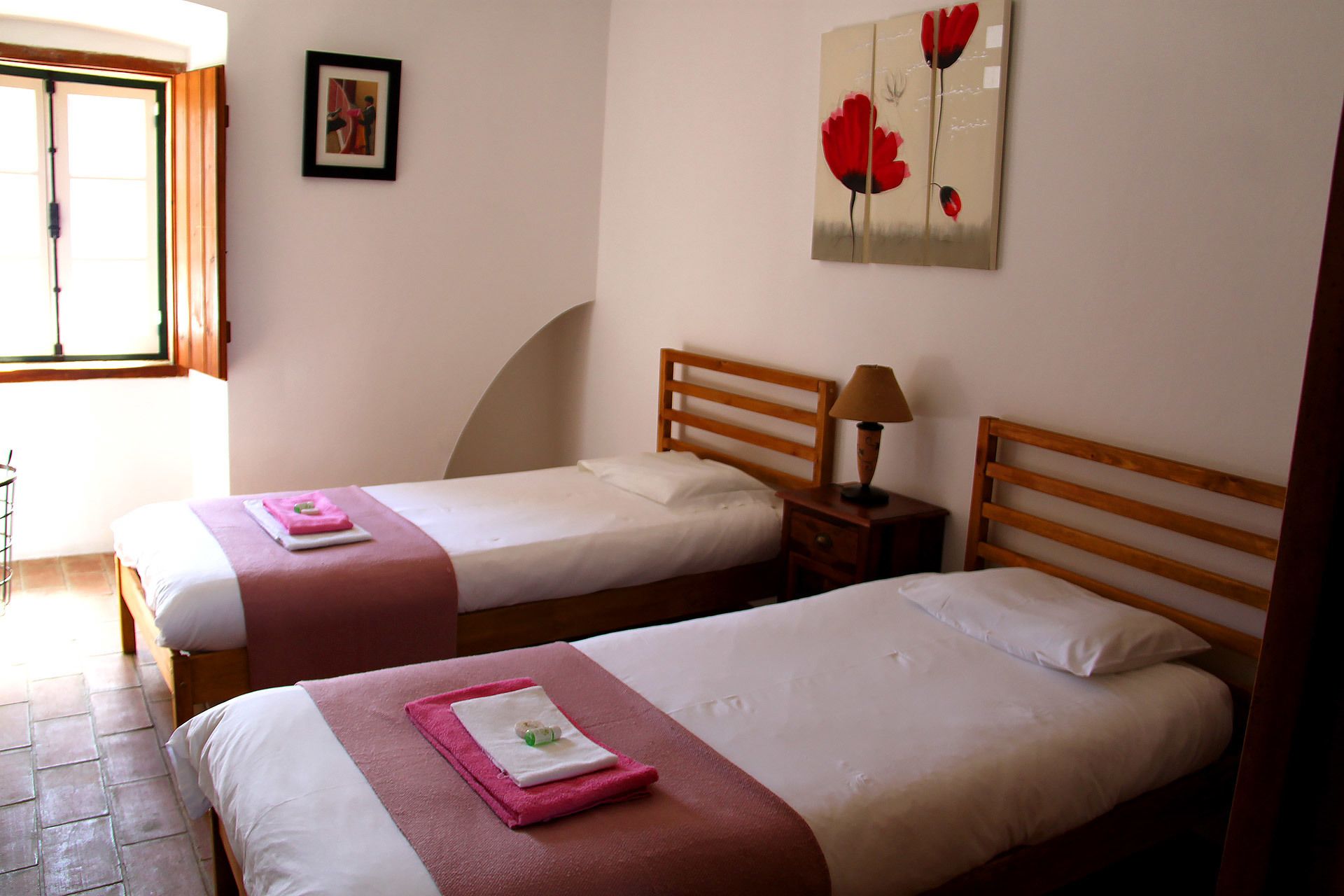 A hotel room with two single beds, each with a wooden headboard, a bedside table with a lamp, and a window with a view.