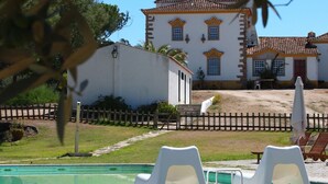 Zona de piscina con sillones blancos, una cerca de madera y un edificio con tejado de tejas al fondo.